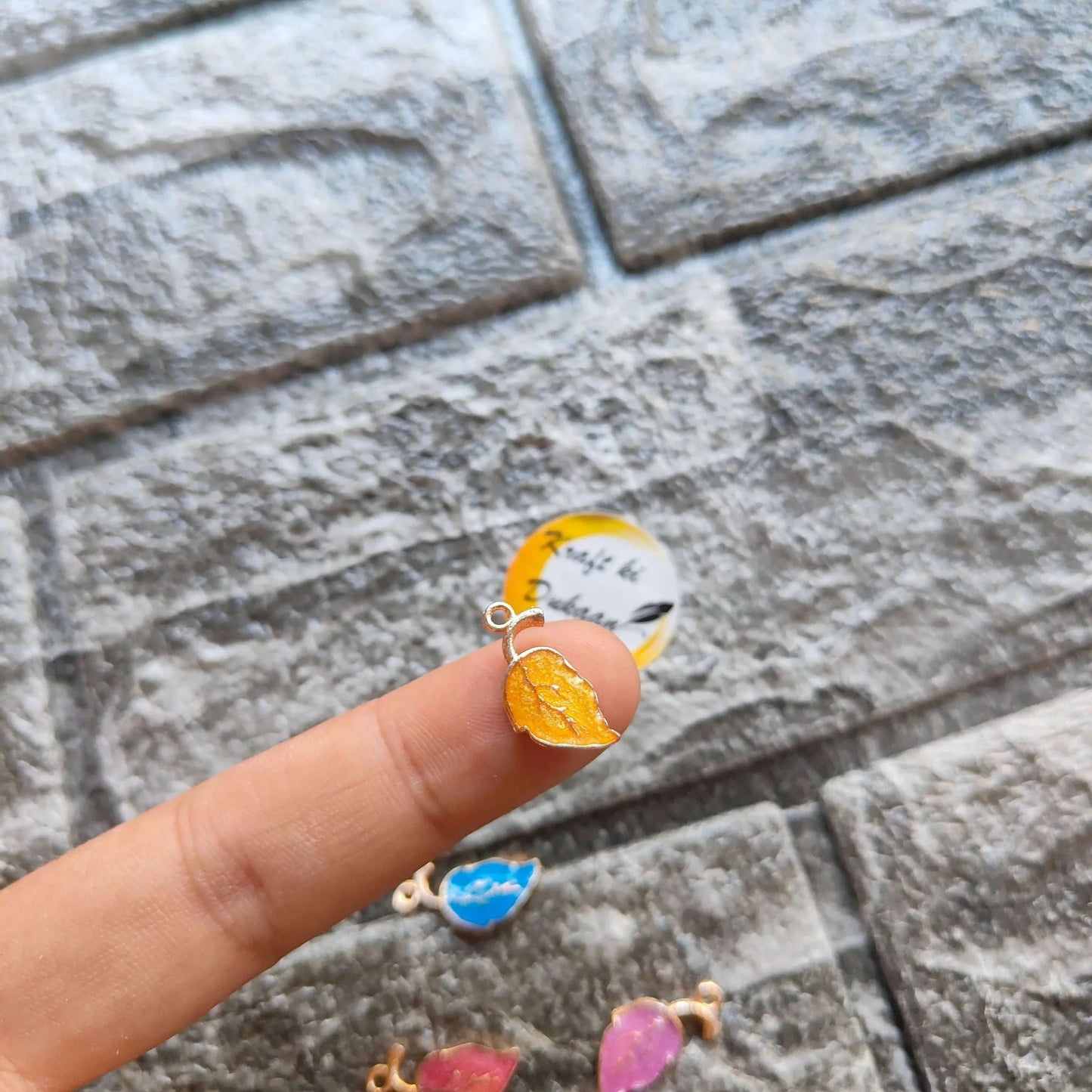 Orange leaf metal charm with gold-toned loop, held above Multi Leaf charms pack