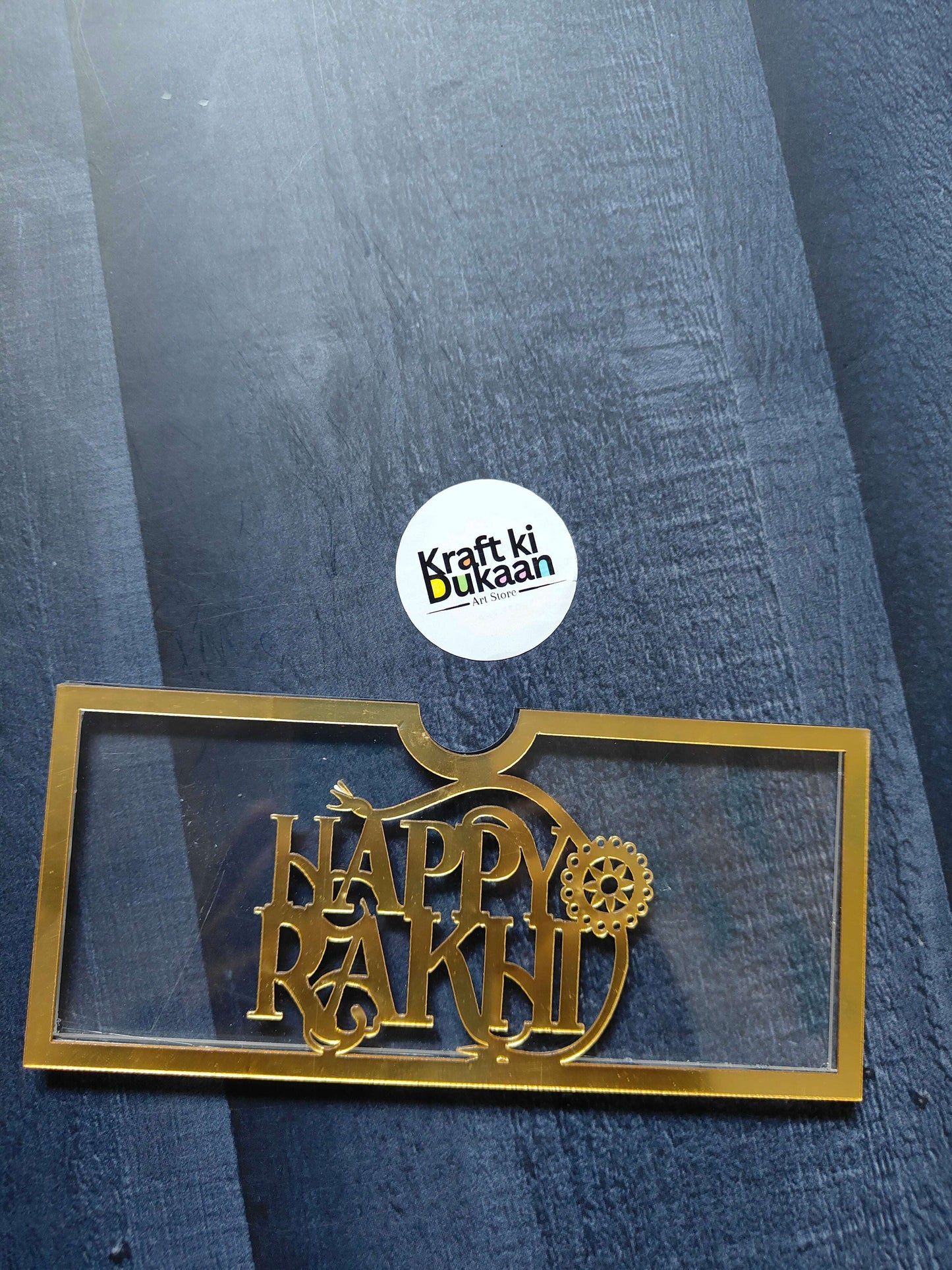 Golden acrylic Rakhi envelope with “HAPPY RAKHI” text and gear-style motif