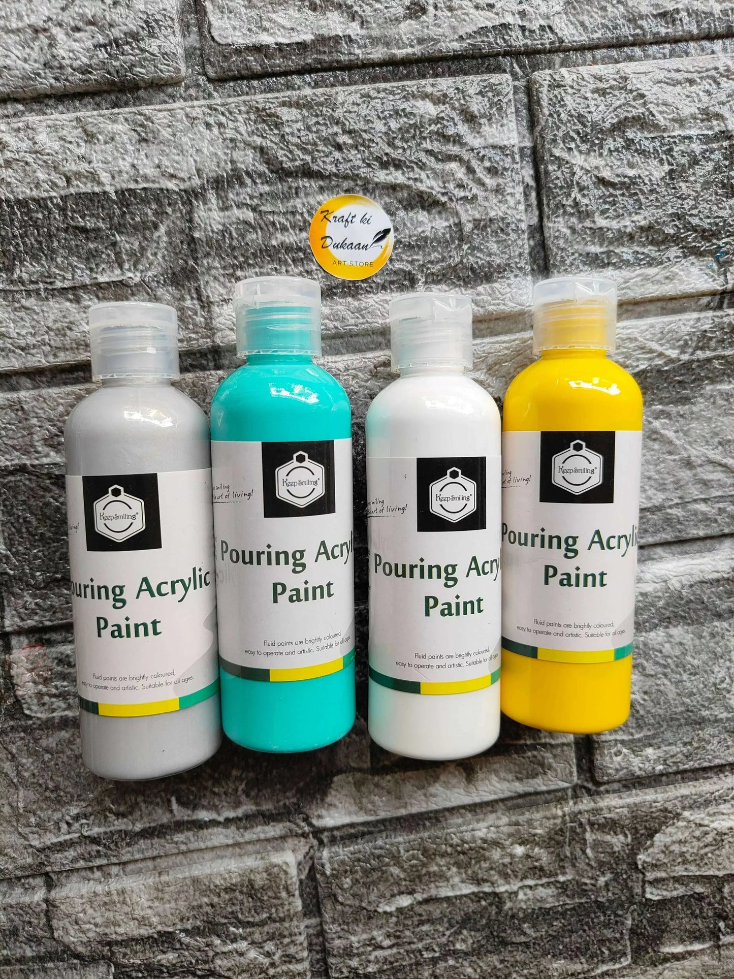 Four acrylic pouring paint bottles: gray, teal, white, and yellow, standing together