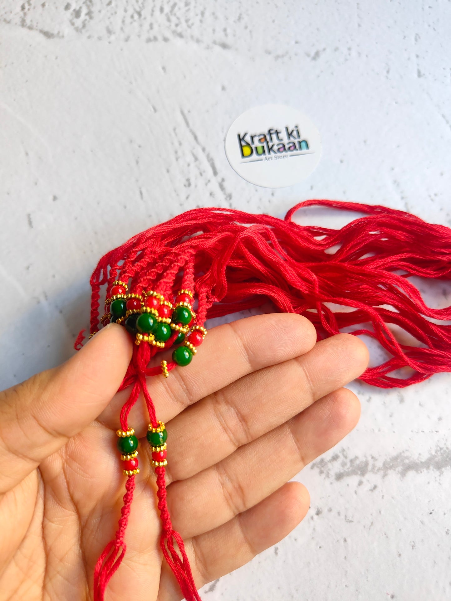 Red rakhi dori in hand, green and gold bead charm with dangling threads