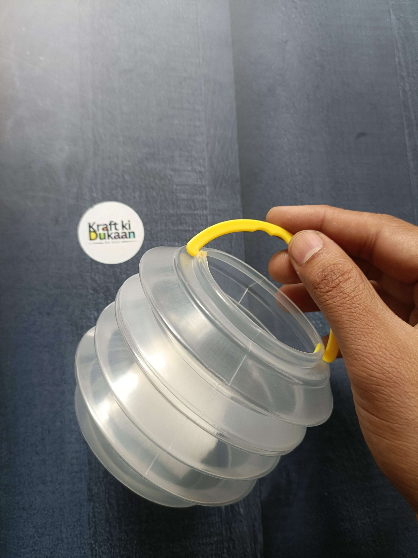 Folded paint brush wash bucket held by hand; clear stacked panels with yellow handle.