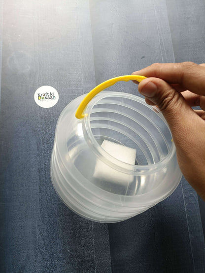 Hand holding folding paint brush wash bucket; yellow handle on clear container.