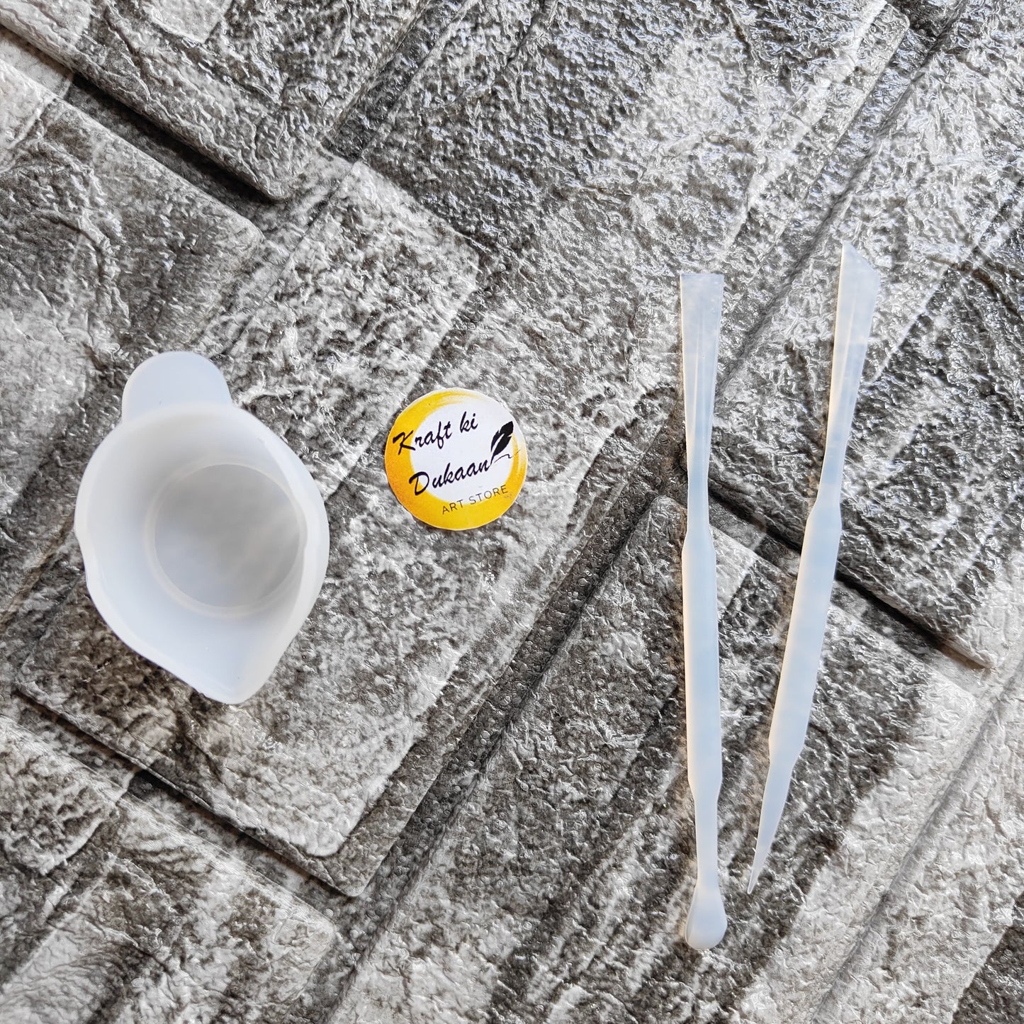 Kraft ki Dukaan resin mixing cup kit with silicone cup and two clear mixing sticks on textured surface