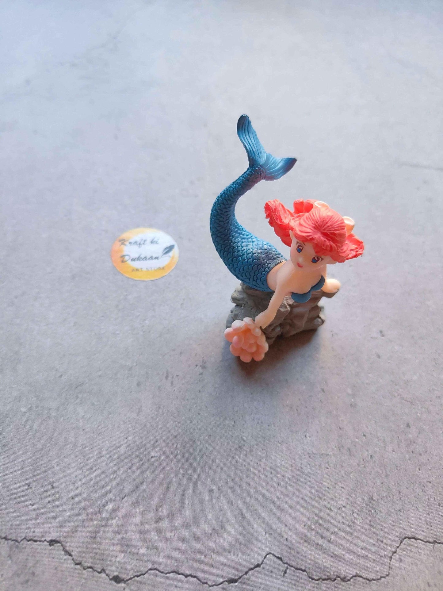 Mini mermaid figurine with blue tail and pink hair, holding orange flower