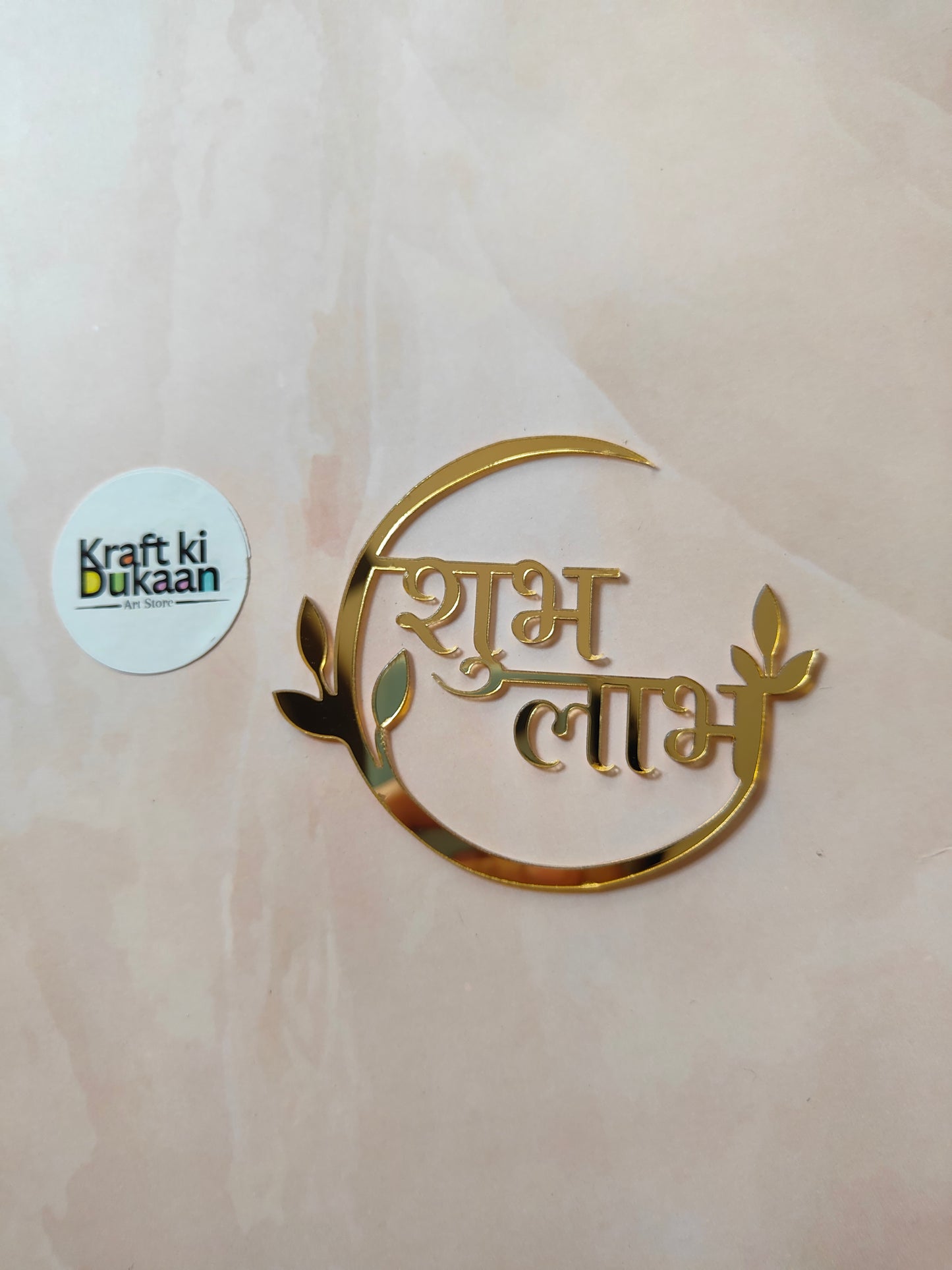 Gold circular acrylic cutout with 'Shubh Labh' Devanagari text and leaf accents