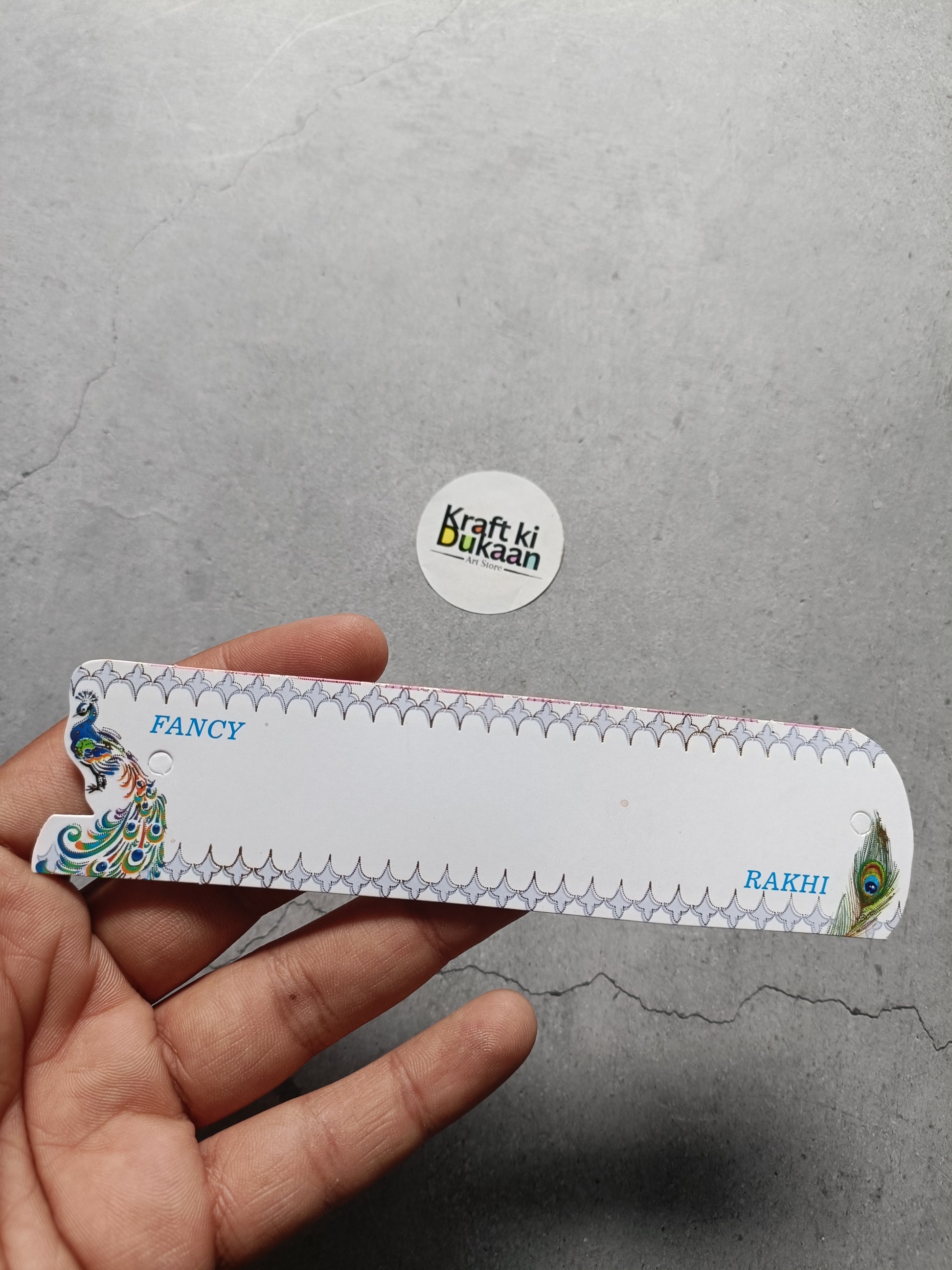 Rakhi greeting card held in hand, white with decorative border and peacock corner art