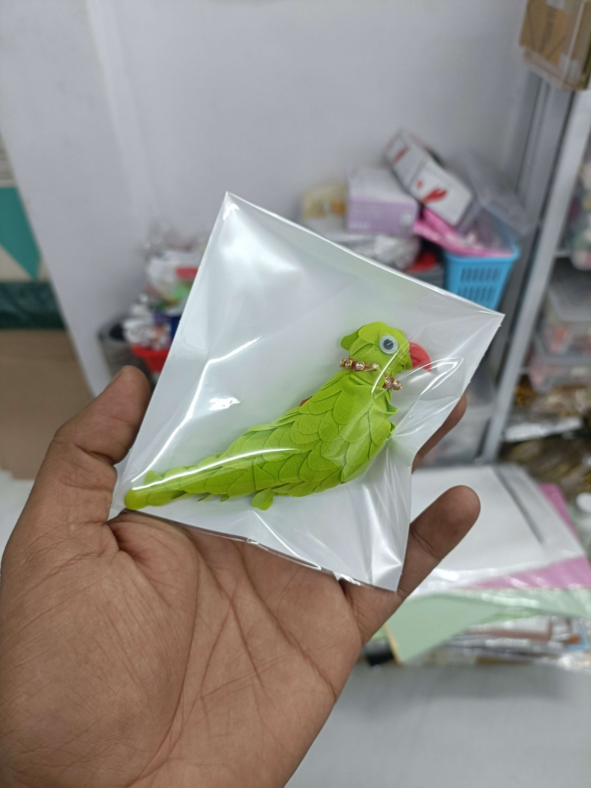 Green parrot-shaped fabric pin with bead eyes, packaged in clear plastic sleeve