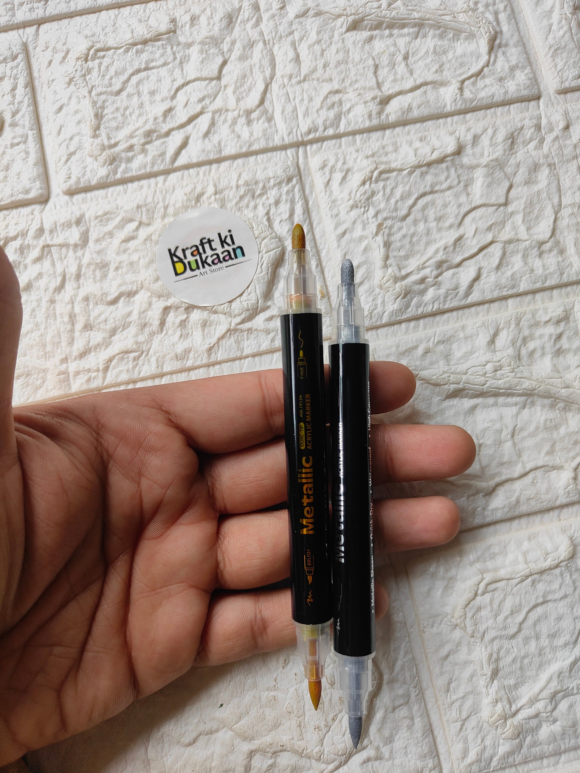 Dual-tip metallic acrylic markers held in hand, gold and silver tips visible